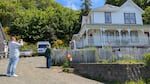 On a sunny day, two people stand by the famous "Goonies House" in Astoria. One holds a camera while the other poses for the picture with the house behind them.