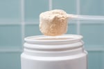 A close-up of a scoop of beige protein powder held above an open white plastic jar, all set against a blue tile wall.
