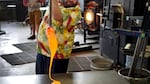 A man in a floral shirt drips molten glass from the end of a rod onto a metal table.