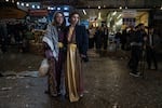 Avjin Yazgan, 21, stands with her mother Pelda Yazgan, 43, are visit Akre to celebrate the holiday. Avjin is wearing a maroon dress and a keffiyeh. She is holding a bag of flatbread. Pelda is wearing a gold and maroon dress with a suit jacket over top. 