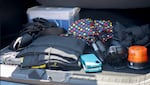 A collection of objects in the trunk of Tea Godfrey's car including a file box, a blanket, an umbrella, a light, two phones, and measuring tools.