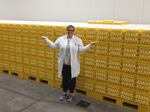 Wearing a white lab coat, dark pants and sneakers, the American Egg Board's Emily Metz stands in front of a multitude of yellow stacked trays that are filled with eggs. She is smiling and is gesturing with her arms stretched out from her sides.