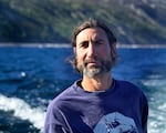 A man looks ahead while pictured with water and a steep hillside behind him.