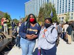 Two young woman wearing facemasks stand facing the camera with several people gather behind them in front of a large office building.