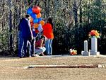People holding colorful balloons stand together around a grave site, as one person kneels down and touches the ground.