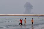 Three boys play in the shallow waters of the Strait of Hormuz, as a plume of smoke rises from an explosion in the background, off Bandar Abbas, Iran, Monday, July 13, 2026.