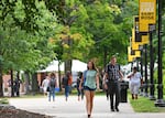 This photo shows an outdoor scene at the College of Saint Rose. Students wearing backpacks are strolling along a walkway in both directions, and leafy trees stand in the background.