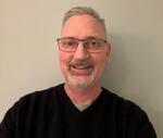 A man with gray hair and a beard smiles while wearing glasses and a black shirt.