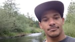 A man wearing a baseball cap looks into the camera. Behind him is a river with grassy banks and trees.