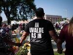 A person wearing a t-shirt that reads "In Uvalde as in Heaven" stands with his back to the camera and holds hands of people standing on either side of him.