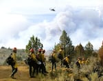 Firefighters head in to construct a fire line in triple-digit heat as a helicopter flies overhead to cool off hotspots with water on the Falls Fire, burning on the Malheur National Forest