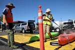 A pair of people wearing boat hats and construction vests stand in a parking lot. One hoses down a plastic kayak.