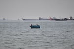 Two men sit in a small boat on the water as a mix of bulk carriers, cargo ships, and service vessels line the horizon in the Strait of Hormuz off Bandar Abbas, Iran, Monday, April 27, 2026.