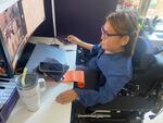 Tabi Haly, a woman in a blue top, sits in her wheelchair in front of a computer screen. She talks to others whose images in boxes on the screen.