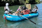 Three people on a blue raft that is full of trash, including a grocery cart, suitcase, buckets, and an old metal sign.