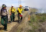 Three people on a gravel road, with one using a drip torch to set a fire.