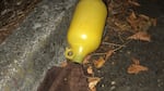 A yellow jar with a brown towel stuffed into its mouth rests amongst leaves on the side of a road.