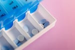 This photo shows a blue and white daily pill container filled with Aleve and Tylenol for each day of the week, on a pink backdrop.