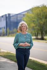 Juli Briskman stands on the sidewalk along the street in front of the Vantage VA2 data center in Sterling. She's wearing blue pants and a teal long-sleeved shirt. The data center is a large blue building in the background.