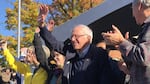 A crowd of people who are waving, clapping and smiling. Light falls on the face of Bernie Sanders, who is in the center of the image.