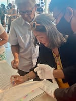 Chisao Hata (center) uses a hanko to stamp next to the names of relatives in an Ireichō while her cousin Luke Iwata (left) looks on.  The Ireichō contains the most comprehensive list of interned Japanese and Japanese Americans to have ever been compiled. 