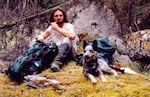 A man sits on a mossy forest floor with a backpack nearby and a dog to his left.