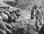 In this black-and-white photo from 1981, Ali Khamenei (on the right side of the frame) speaks to members of Iran's armed forces, who are wearing helmets and are seated or crouched in rows on the ground on the left side of the frame.