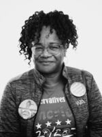 This black-and-white photo is a portrait of Sandi Lytle from about the waist up. She's wearing a shirt that says "Conservatives" and "America" on it. Over the shirt she's wearing an unzipped jacket with two buttons pinned to the front of it. One button says "Make America Great Again," and the other button shows a photo of a woman wearing a basketball jersey.