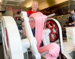 A batch of pink taffy is pulled in a century-old machine. Behind the action, a white man with a bald head wearing a read shirt and black apron smiles.