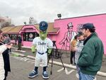 Dillon the Pickle, mascot for the Portland Pickles, and staff of the baseball team greet members of the press outside a pink doughnut shop on a cloudy morning.