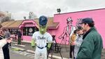 Dillon the Pickle, mascot for the Portland Pickles, and staff of the baseball team greet members of the press outside a pink doughnut shop on a cloudy morning.