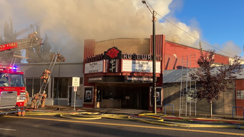 A theater covered in smoke while firefighters climb a ladder onto a nearby roof.