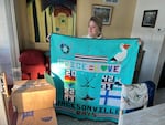 A women holds up a light blue quilt with symbols like a turtle, hockey sticks and a pelican sewn on it.