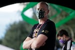 A bald man stands with his arms folded wearing a green mask as he stares into the camera.