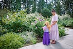 A young girl and woman admire pink roses, with a backdrop of tall evergreen trees.