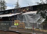 Construction workers work on the outside of a building behind a chain link fence and nearby fir trees.