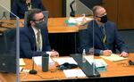 Two men in suits and wearing face masks sit at a wooden table, covered in paperwork, notepads and with microphones. They're separated by Plexiglas barriers.