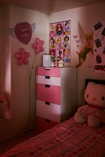 A sliver of light shows a bedroom with a pink dresser, a poster of Disney princesses and Tinkerbell on the wall. There is a large Hello Kitty stuffed animal on a pink bedspread.