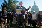 Senator Ted Cruz, wearing a suit and sunglasses, stands outdoors behind a lectern equipped with microphones as he speaks about the Protect College Sports Act at a news conference with NCAA athletes and coaches on September 14 in Washington, D.C. With him are Senator Maria Cantwell and Senator Shelley Moore Capito, who are standing off to the sides of the frame.