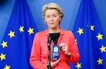 A person standing in front of European Union flags holds up a smart phone to display a digital COVID-19 travel certificate on the screen.