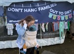 Natalie Kiyah holds her baby in a carrier strapped to her chest and kisses the baby on the forehead while holding back some hair. She stands in front of a white tarp tent that has a blue sign in front of it. The sign says, "Don't hang families out to dry. #FundChildCareNow." There are baby onesies strung up on and around the sign.