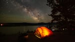 A tent glows with light from within on a dark night. The stars of the Milky War are visible overheard.