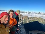 Three people in jackets and winter hats smile at the camera from atop a mountain ridge.
