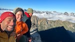 Three people in jackets and winter hats smile at the camera from atop a mountain ridge.