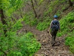 A man wearing a backpack walks a trail with trail keeping hand tools.