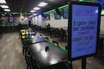 A bilingual white board sign reads, "Welcome. Espere para ser sentado. Gracias/Wait to be seated. Thank you." The sign stands in front of a mostly empty restaurant and bar with TV monitors lining the walls.