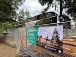 Construction workers work on the outside of a building behind a chain link fence and nearby fir trees.