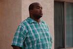 A portrait of DeForrest, who is wearing a green and blue plaid button down shirt, standing outside his home.