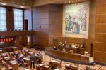 A large government public room with a mural on the wall and people sitting at wooden desks.