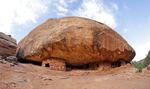 A giant rock with dwellings underneath with blue sky above.